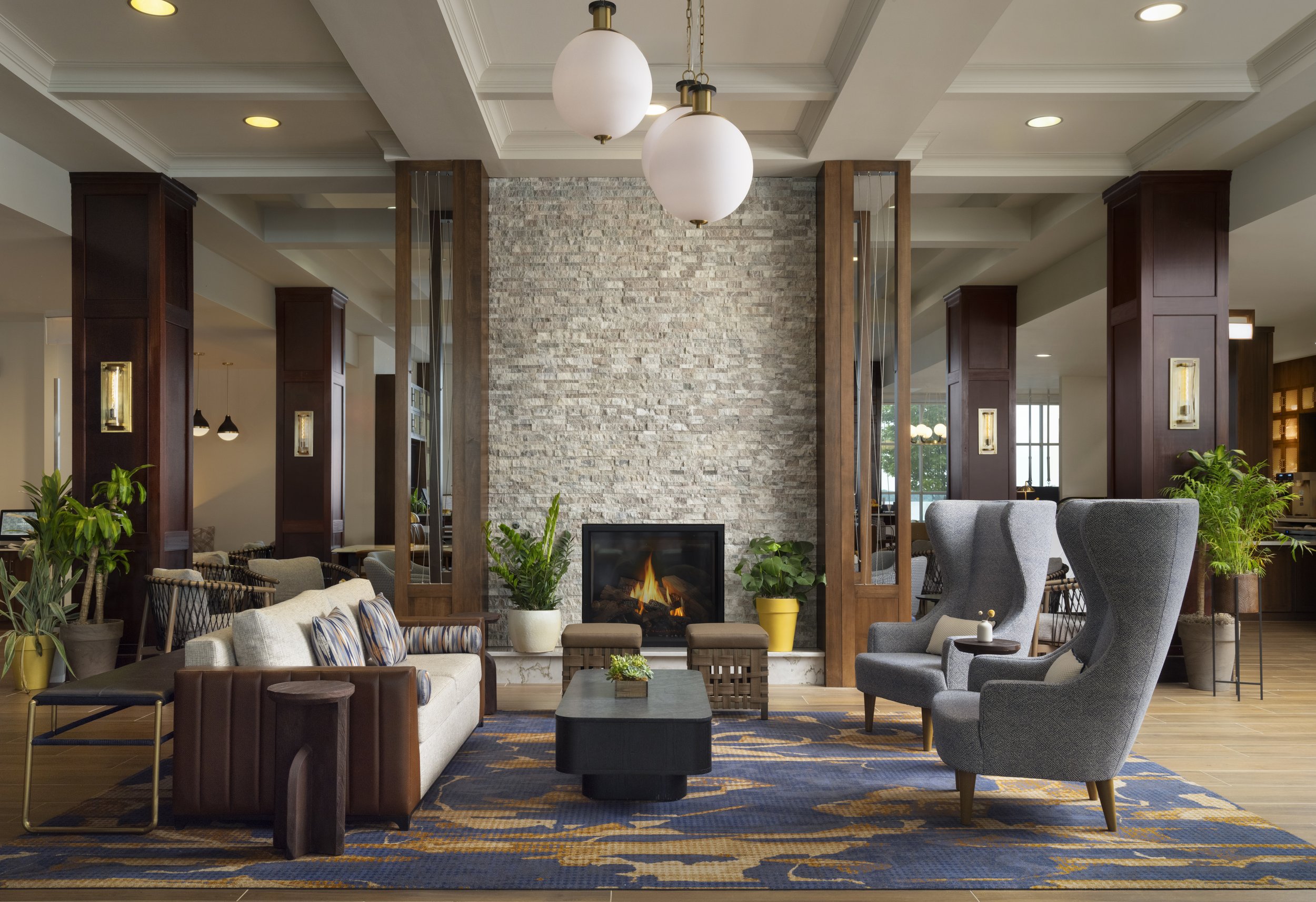 Hotel lobby with a cozy fireplace, beige sofa, two gray wingback chairs, wooden accents, and potted plants.