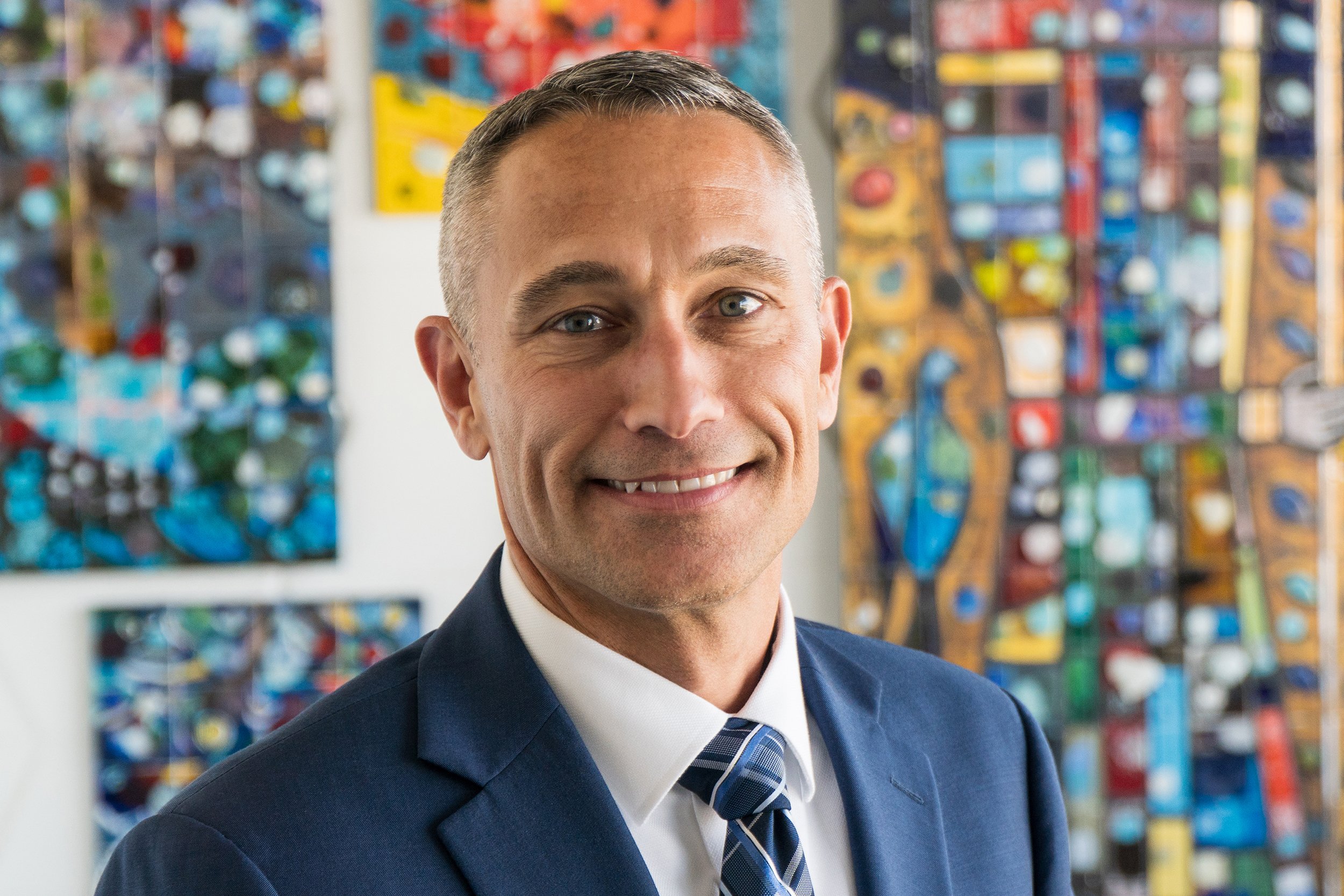 A portrait of Aaron Wilt, Federal Sector, wearing a blue suit with abstract colorful artwork in the background.
