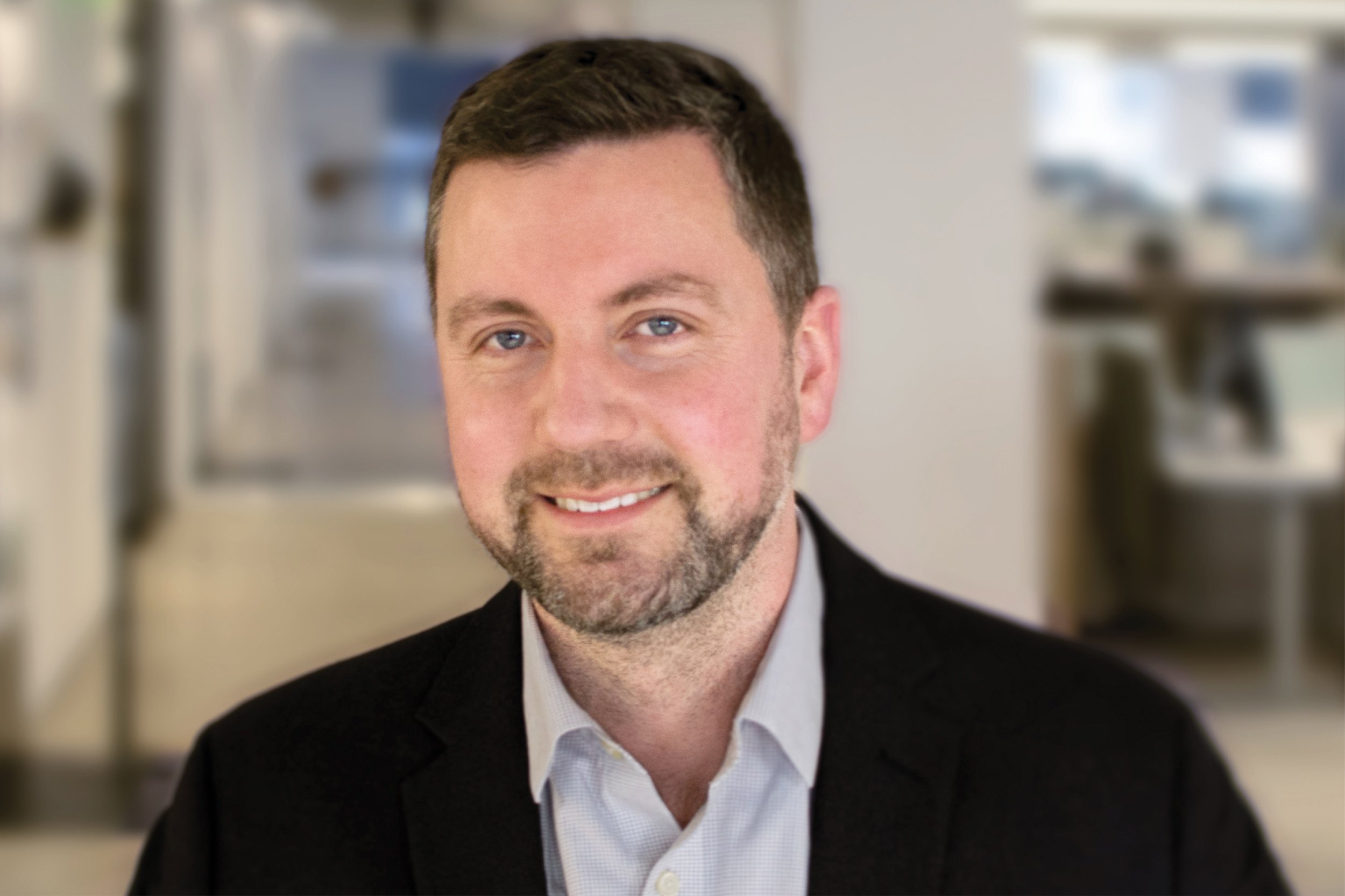 Portrait of Dan Yudchitz, Design Principal, wearing a black suit jacket and white shirt, smiling in an indoor setting with blurred background.