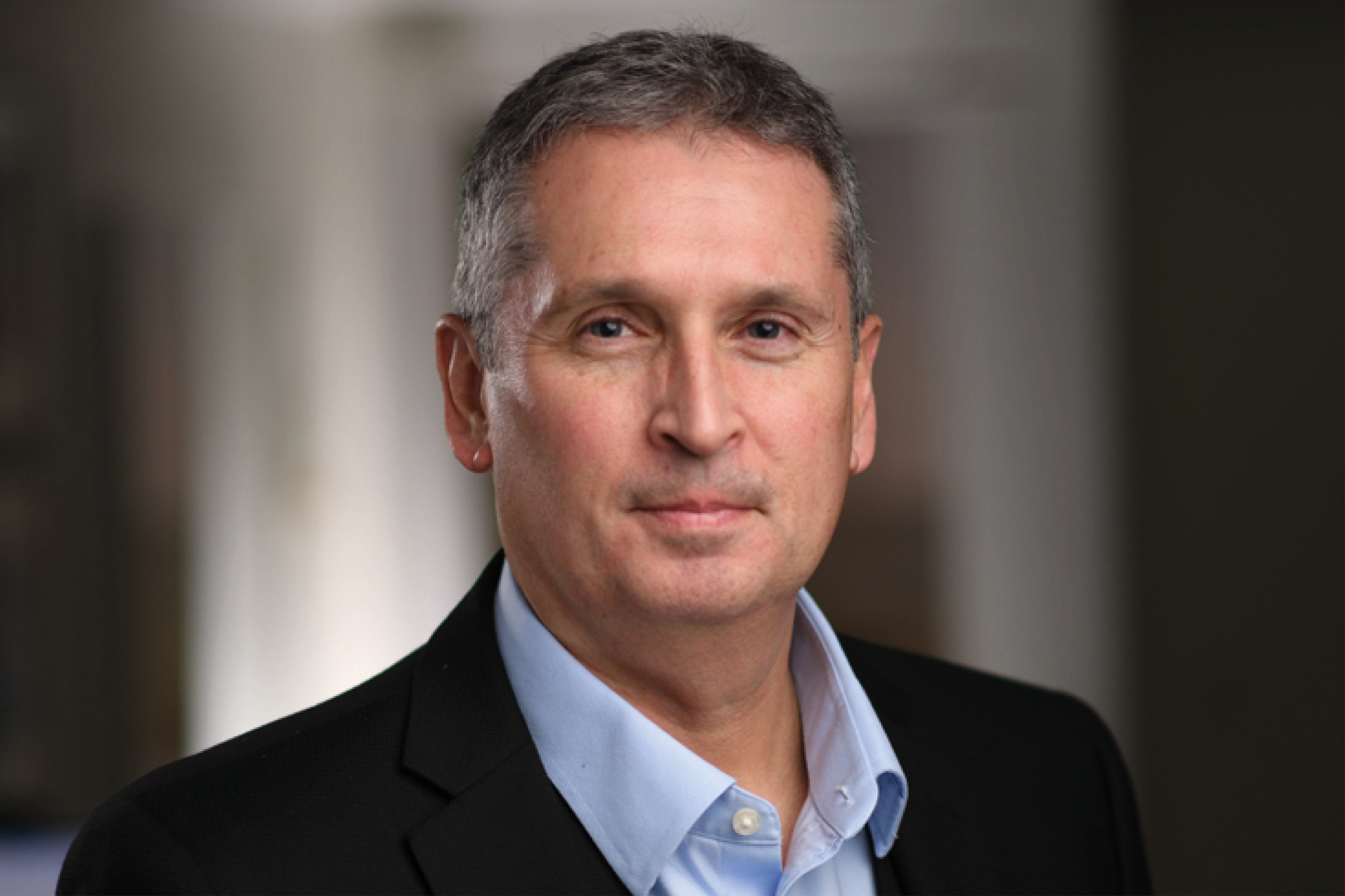 Portrait of a middle-aged man with short gray hair, wearing a dark suit jacket and a light blue dress shirt, standing indoors against a blurred background.