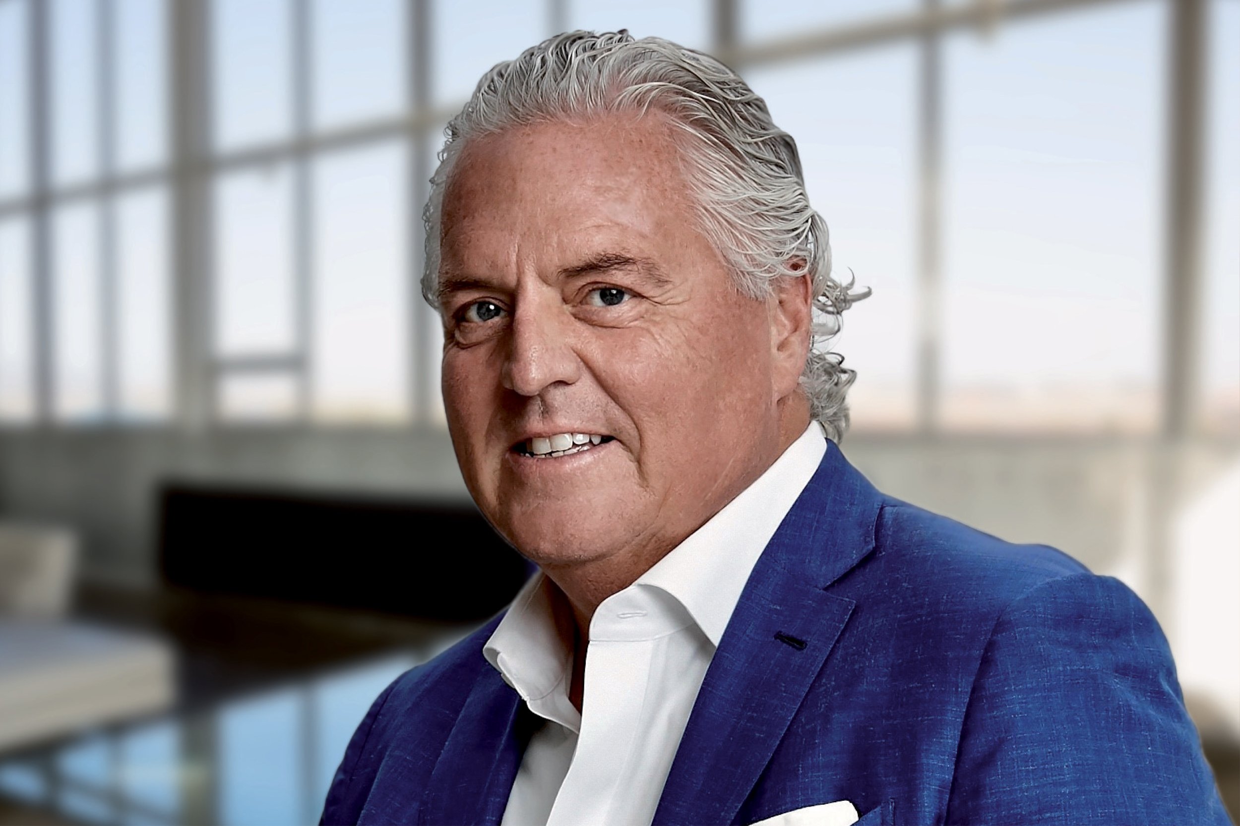 Portrait of Mark Pratt, Hospitality Director, wearing a blue suit and white shirt, standing in a modern office with large windows in the background.