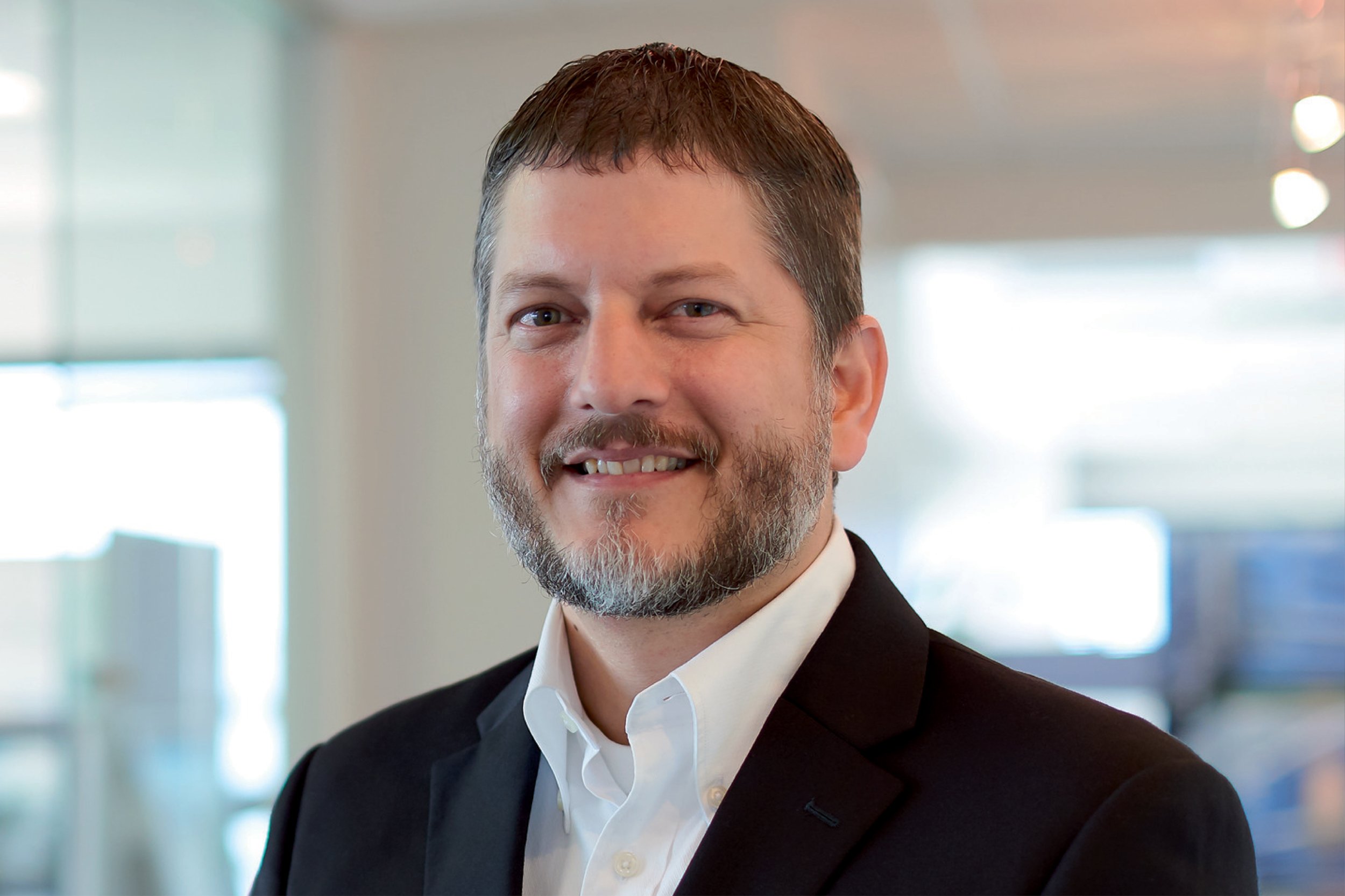 Professional headshot of a smiling man with short brown hair, blue eyes, and a beard, wearing a black suit jacket and white dress shirt, in an office or corporate environment.