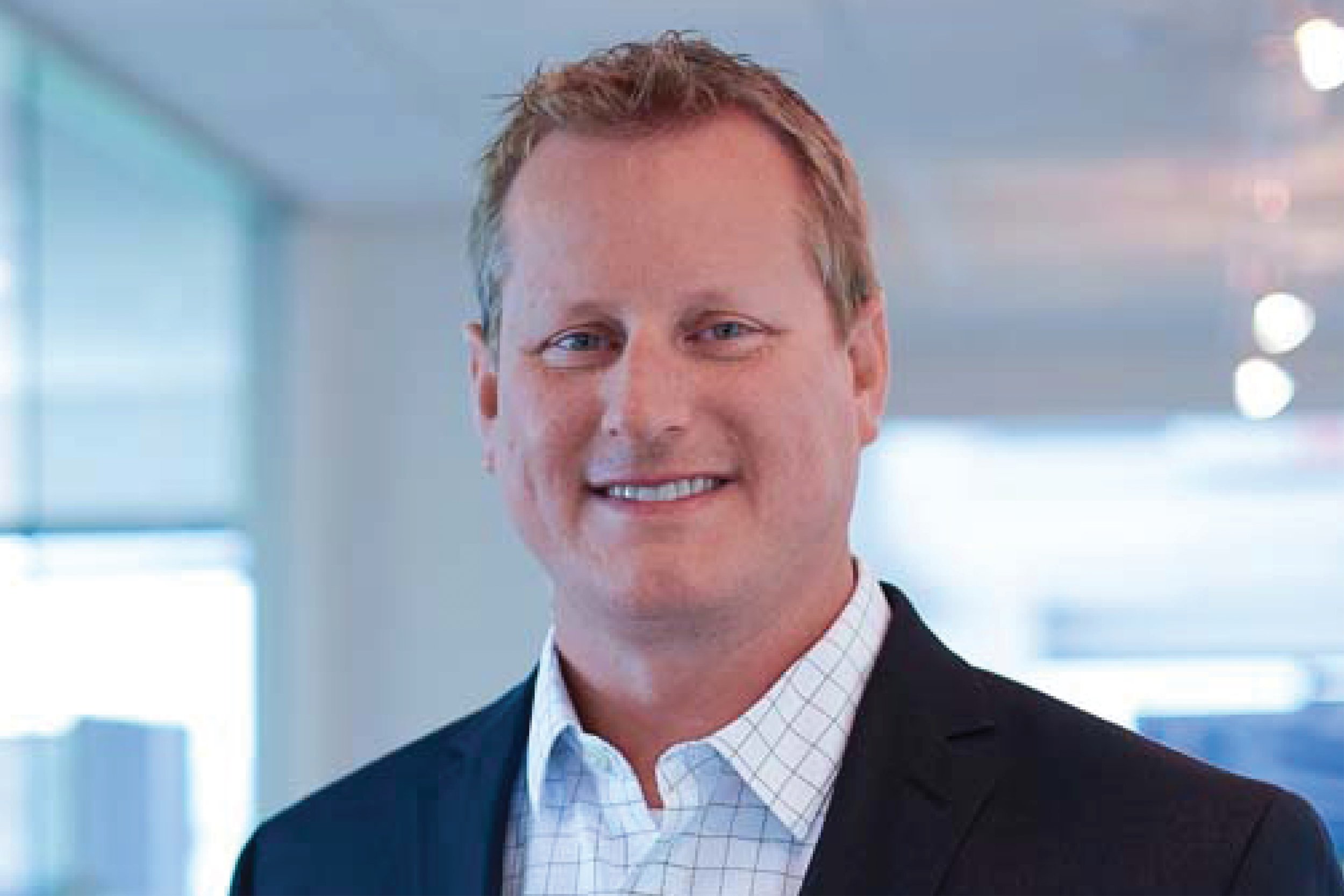 A portrait of a smiling man in a business suit and collared shirt, standing indoors in a bright office environment.