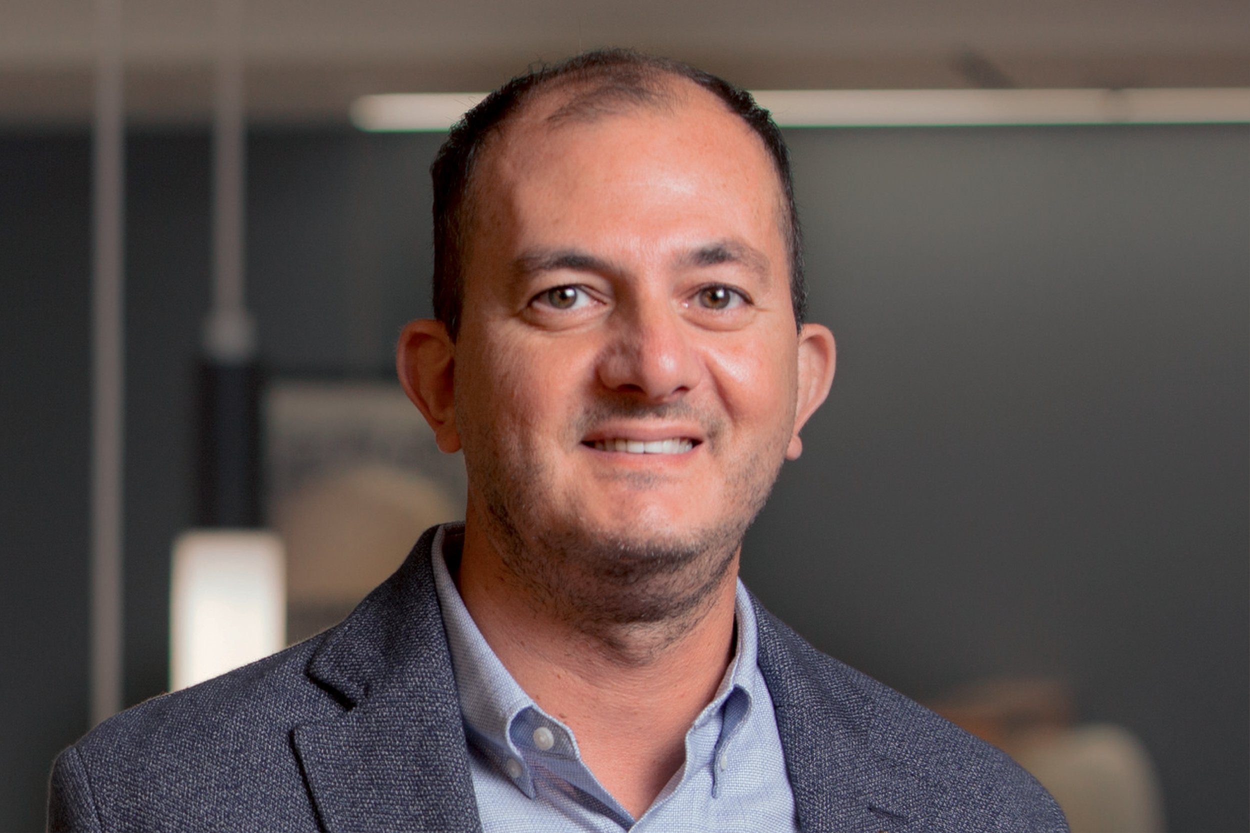 Portrait of Nicos Katsellis, Vice President, wearing a gray blazer and light blue collared shirt, smiling in an indoor office setting.