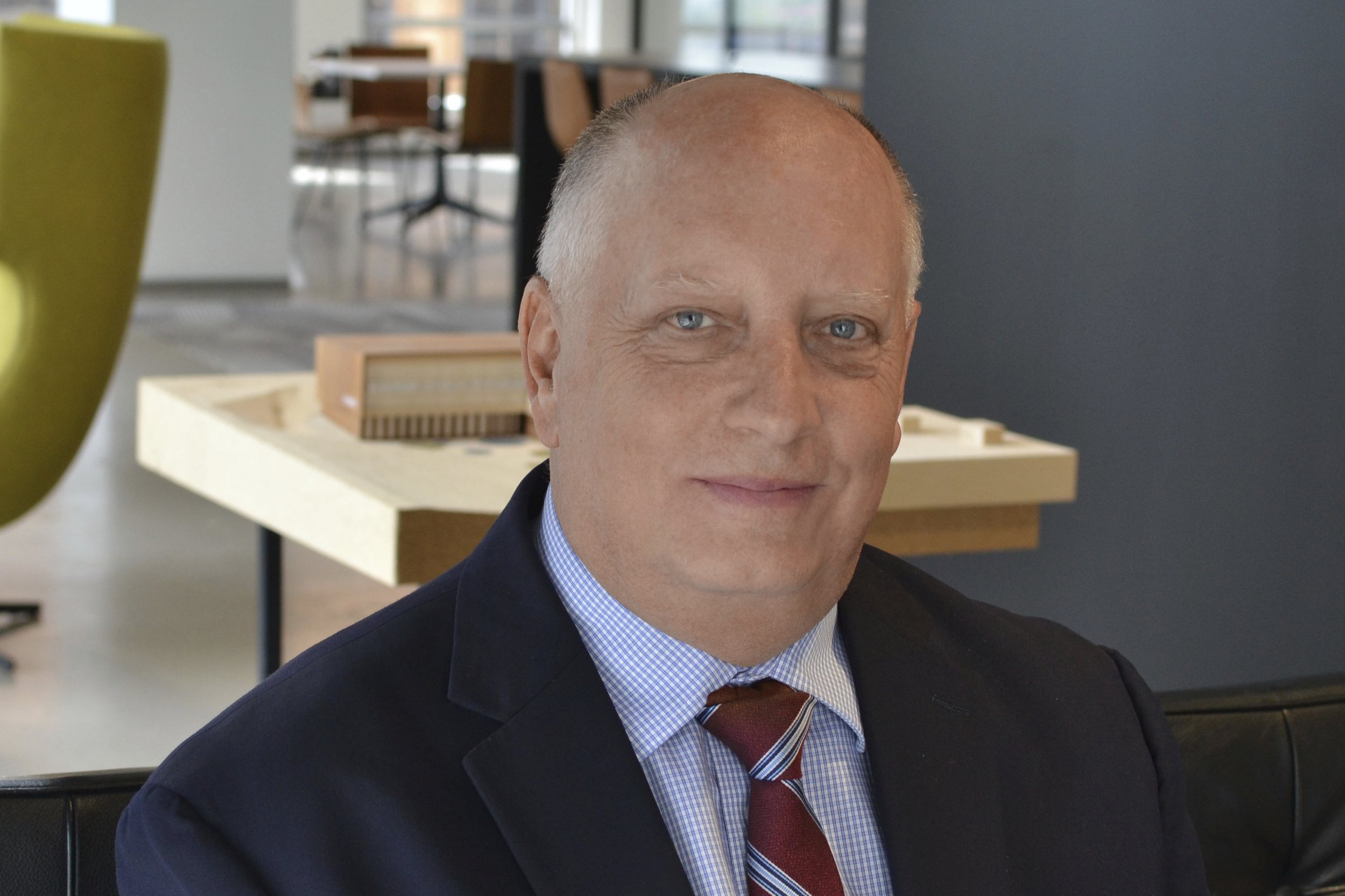 A middle-aged man with a bald head, blue eyes, and light skin, dressed in a navy blazer, blue checkered shirt, and a red striped tie, sitting in a modern office space with a blurred background of wooden desks and chairs.