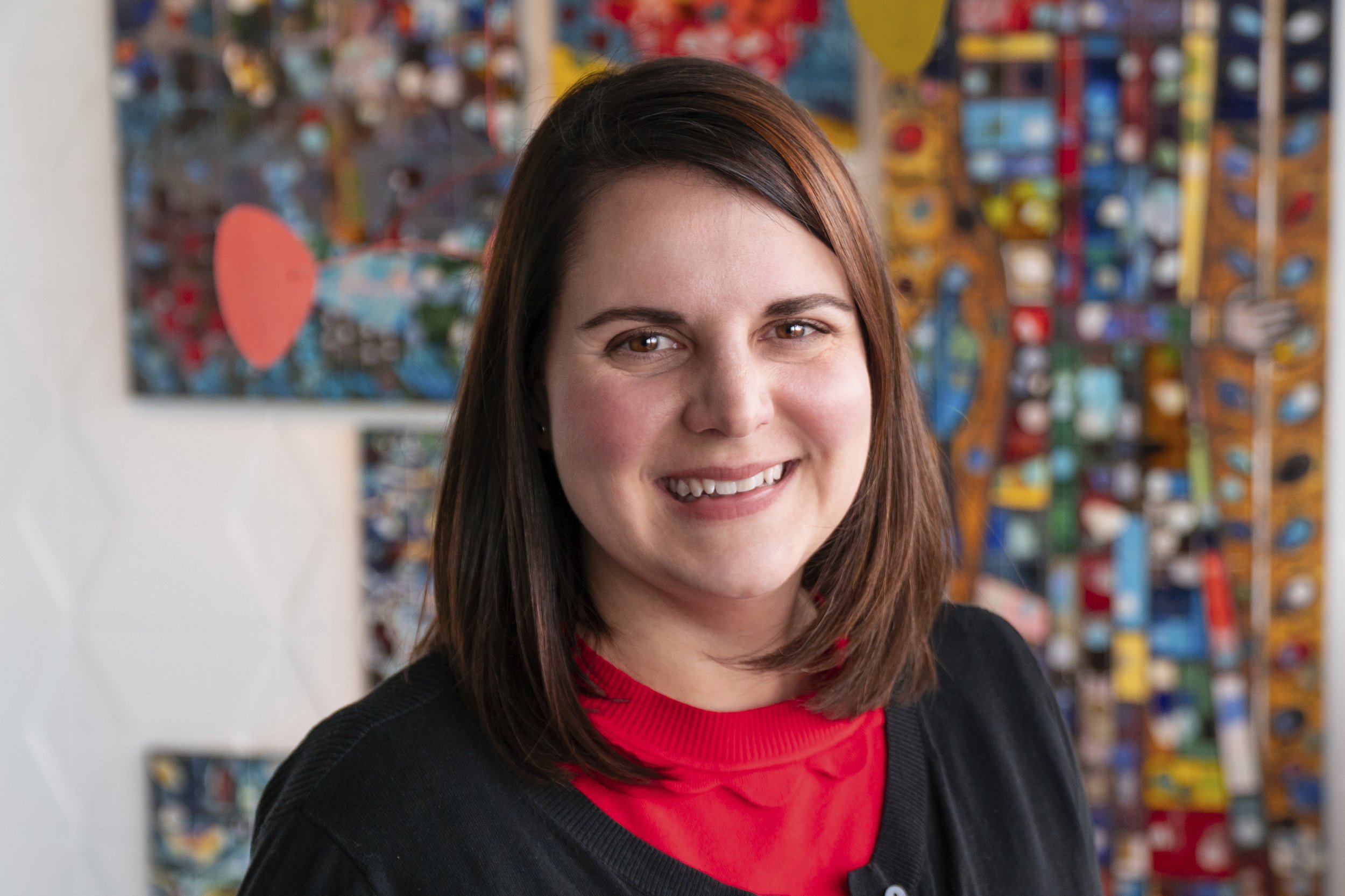 Portrait of Kim Cowman, Vice President, wearing a red shirt and black cardigan, standing in front of colorful abstract artwork.