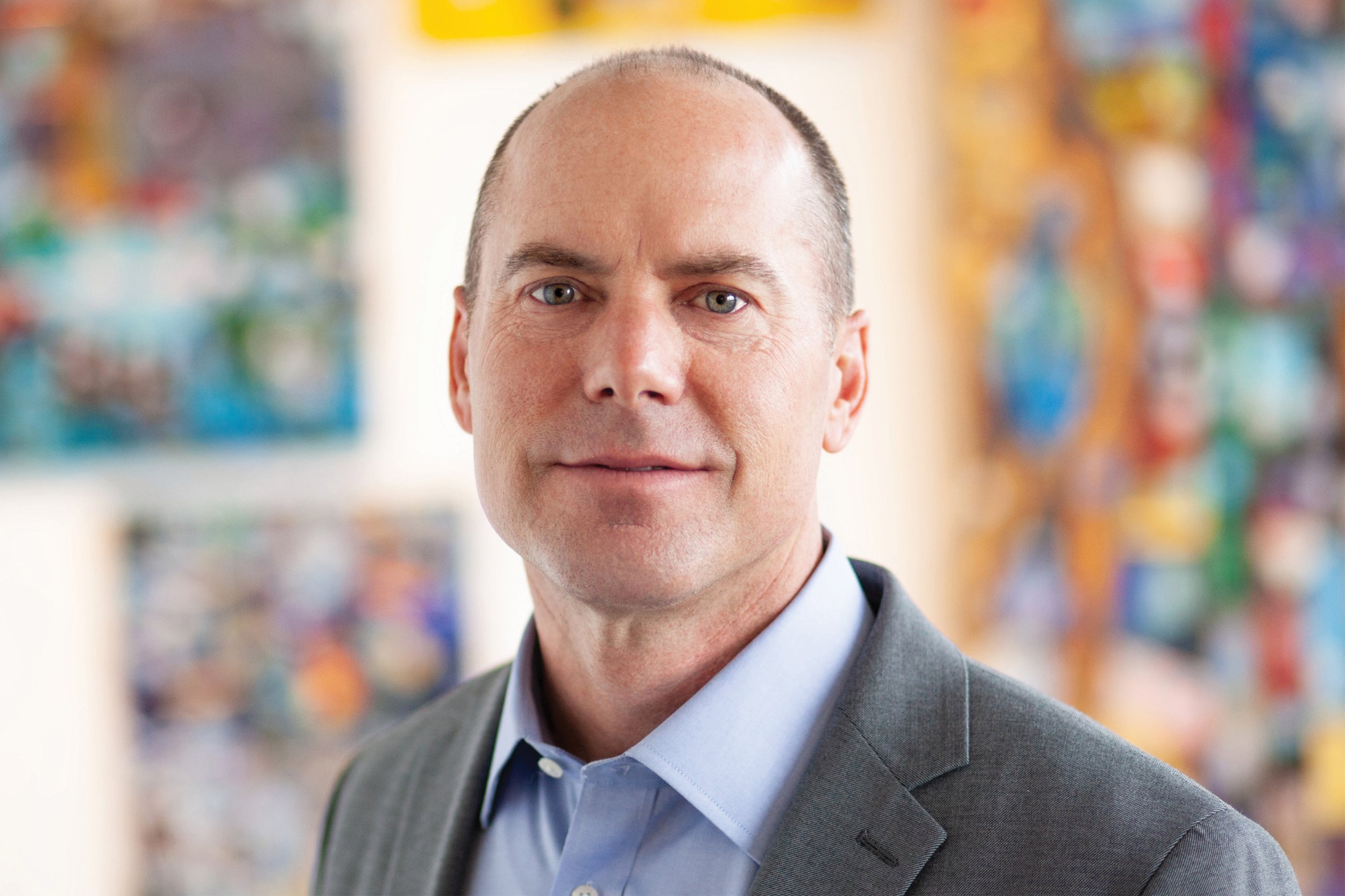 A man in a suit with a blue shirt standing in an indoor space with colorful artwork in the background.