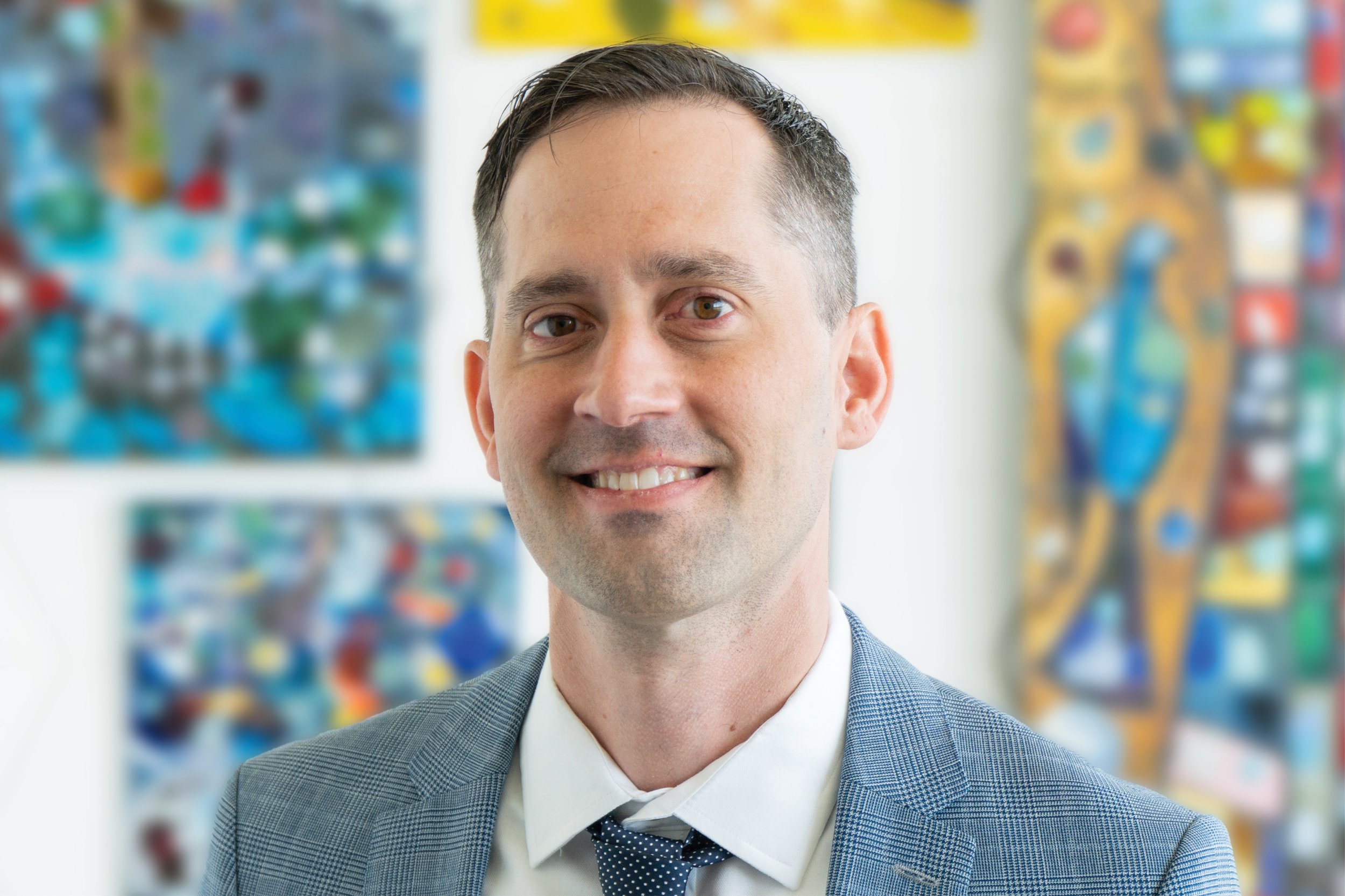 Portrait of a smiling man in a business suit standing in front of colorful abstract artwork in an office setting.